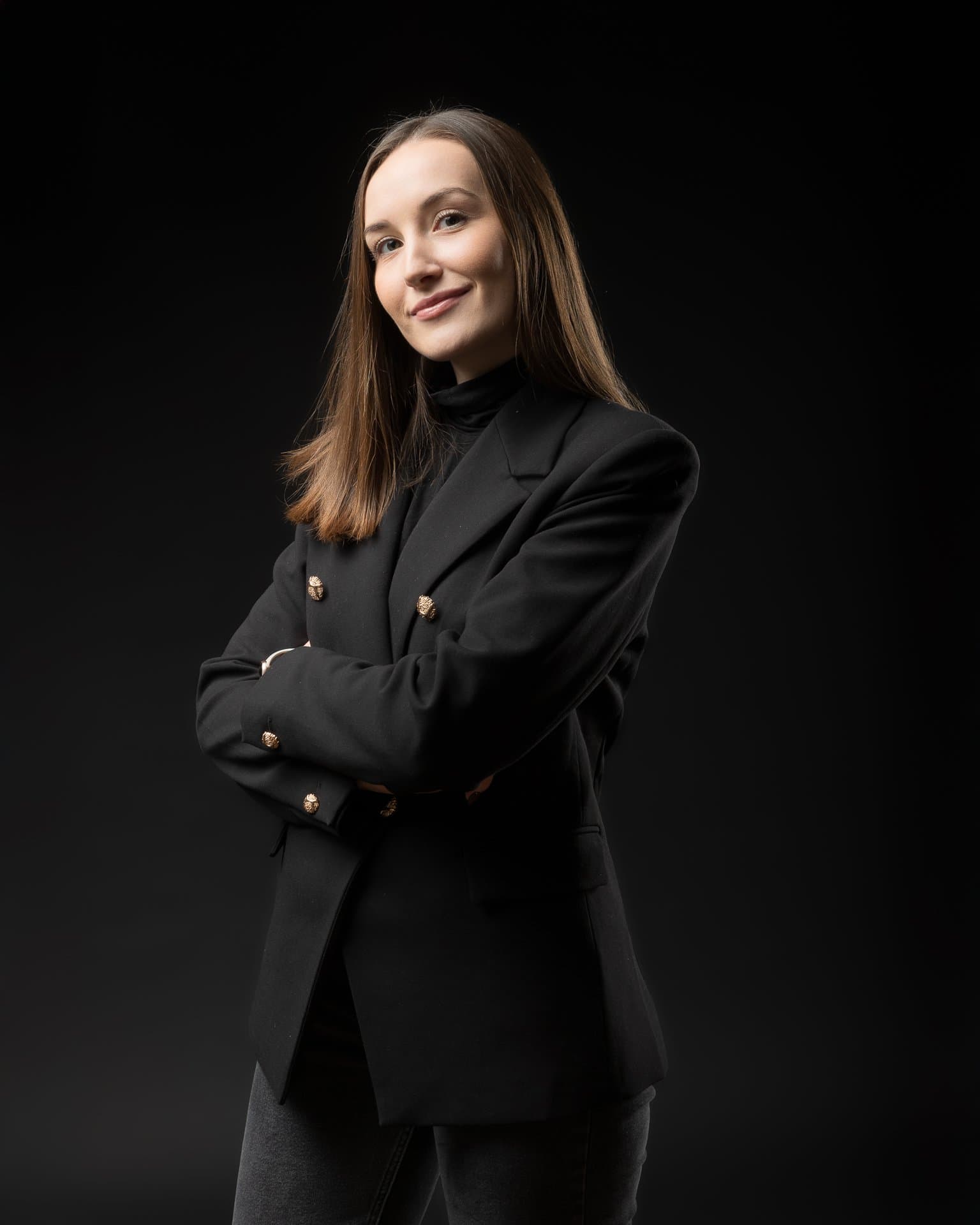 Portret kobiety w czarnej marynarce ze złotymi guzikami, ręce skrzyżowane, czarne tło