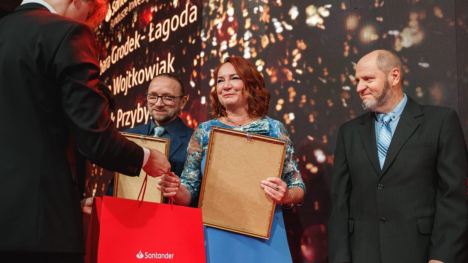 Kobieta i dwaj mężczyźni odbierają oprawione dyplomy na scenie gali, w tle świetlny ekran