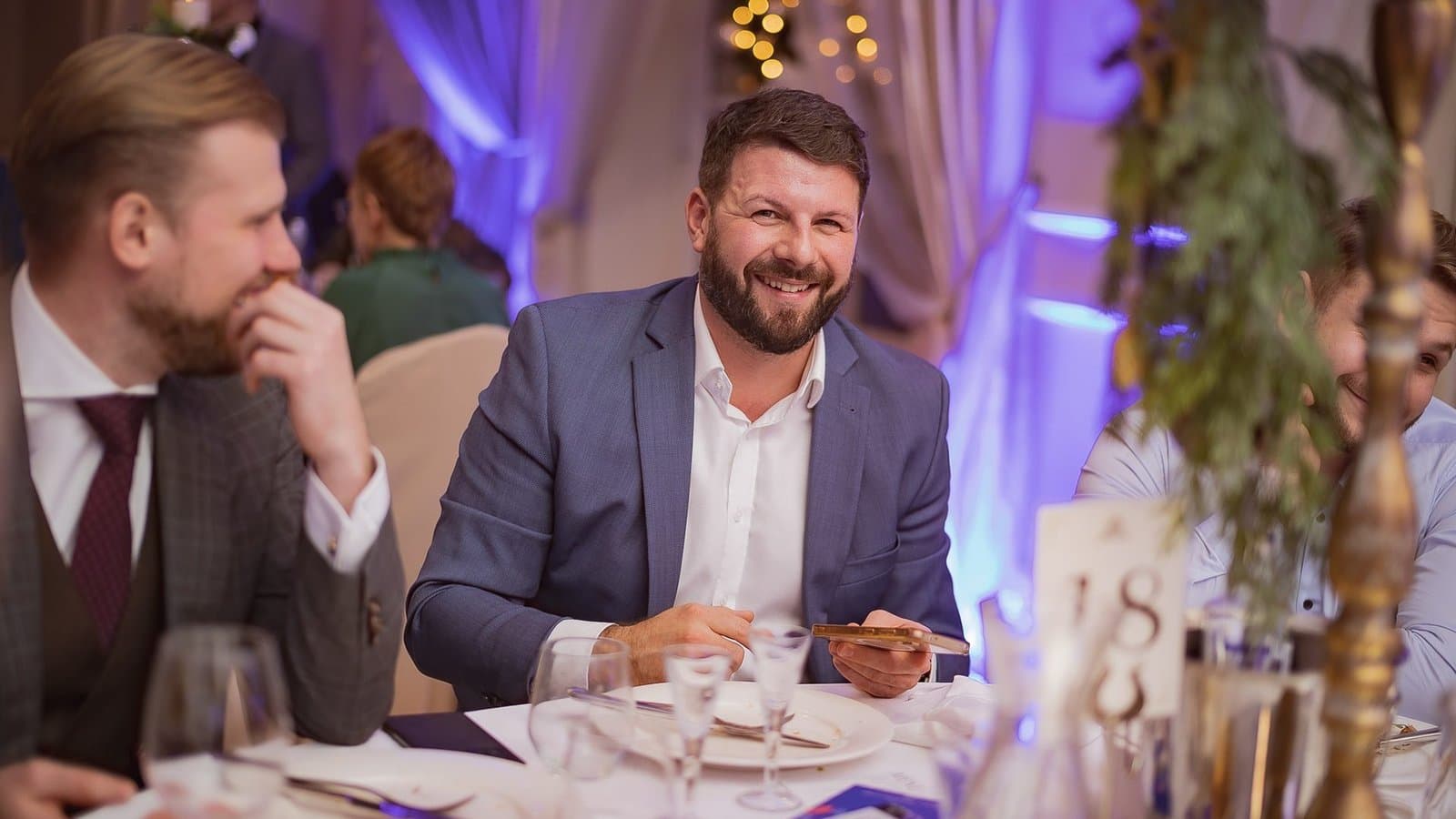 Goście w garniturach śmieją się przy stole podczas wieczornej gali, w tle fioletowe światła
