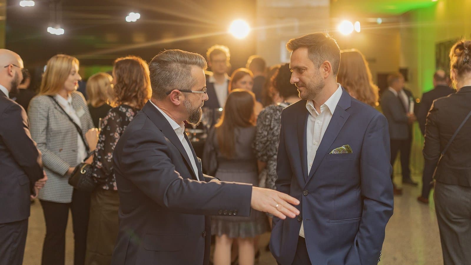 Dwaj mężczyźni w garniturach rozmawiają podczas networkingu, w tle goście i światła sali