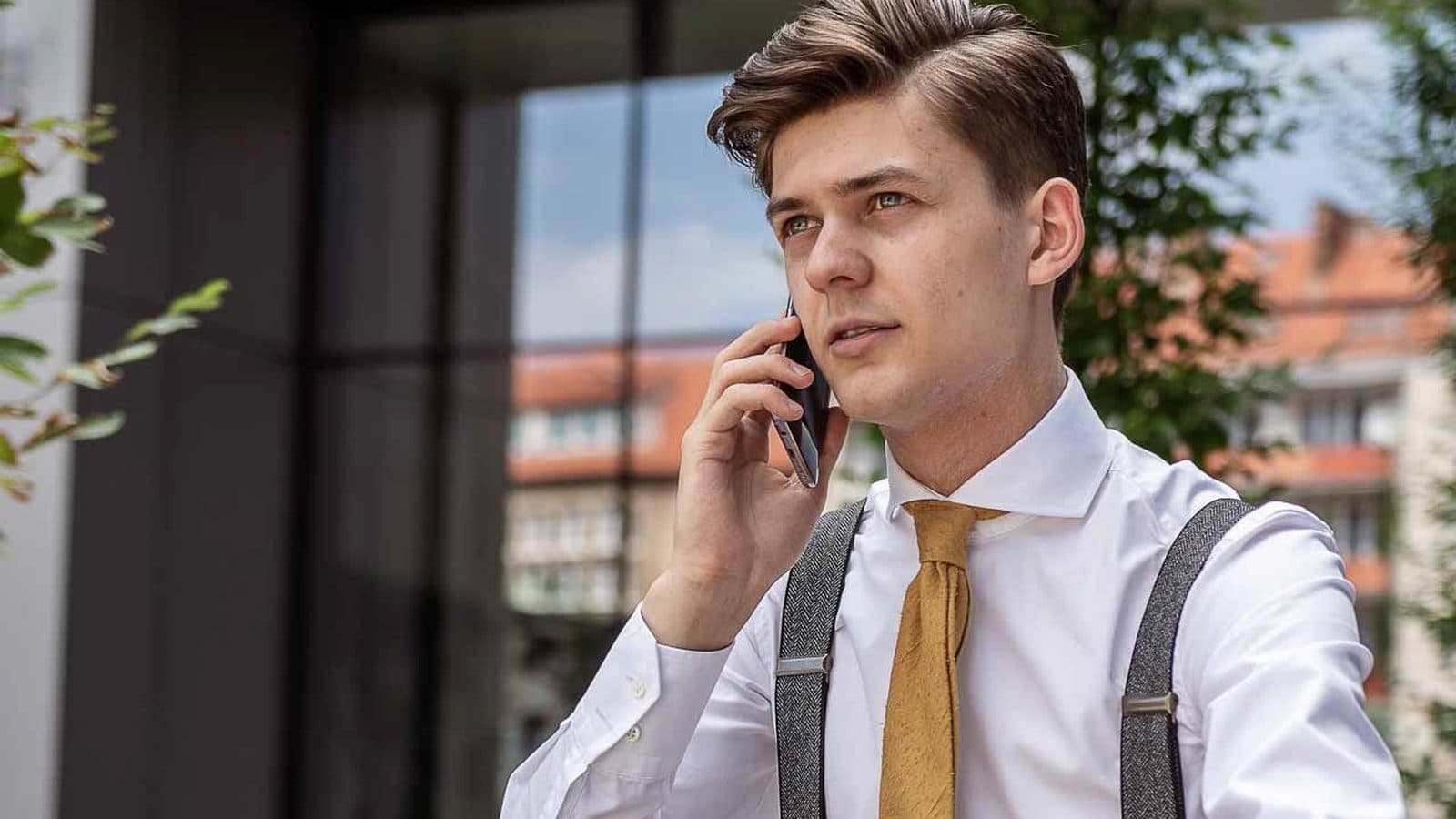 Portret mężczyzny w koszuli i szelkach rozmawiającego przez telefon przed przeszkloną elewacją